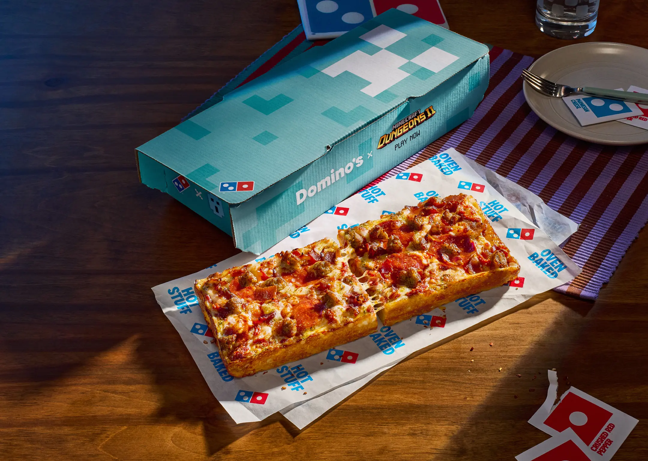A Domino's Minecraft Dungeons II pizza box on a wooden table next to a game controller, with a Domino on branded paper in front of it.